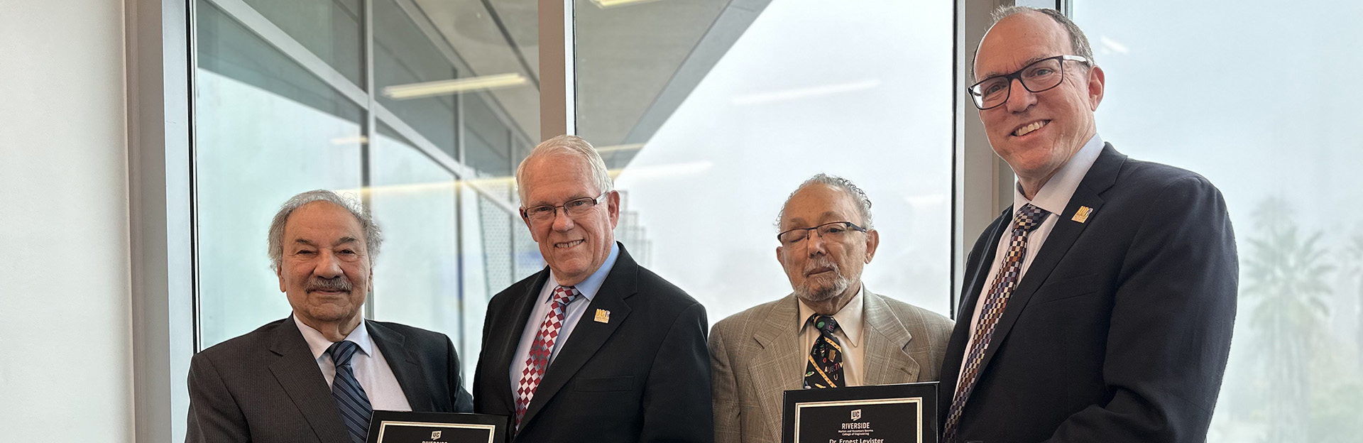 Four men wearing suits and ties are standing side by side, looking at the camera and smiling. From left to right:  Reza Abbaschian and Gordon Bourns hold a plaque, followed by Ernest Levister and Chris Lynch, also holding a plaque. The background features large windows with an outdoor view of trees and what appears to be a building in the distance.