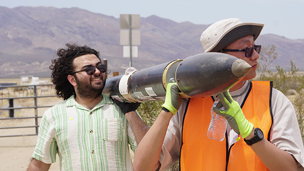 Two engineering students outdoors in the Mojave desert carry a rocket on their shoulders.