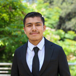 Joe Jimenez, an early twenties Hispanic male wearing black suit and tie with a white shirt smiling