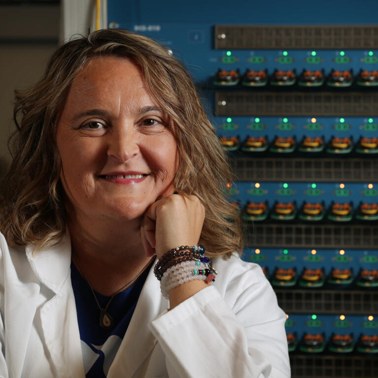 A woman wearing a white laboratory coat smiles at the camera while seated in front of rows of illuminated scientific equipment and electronic testing devices in a research laboratory.