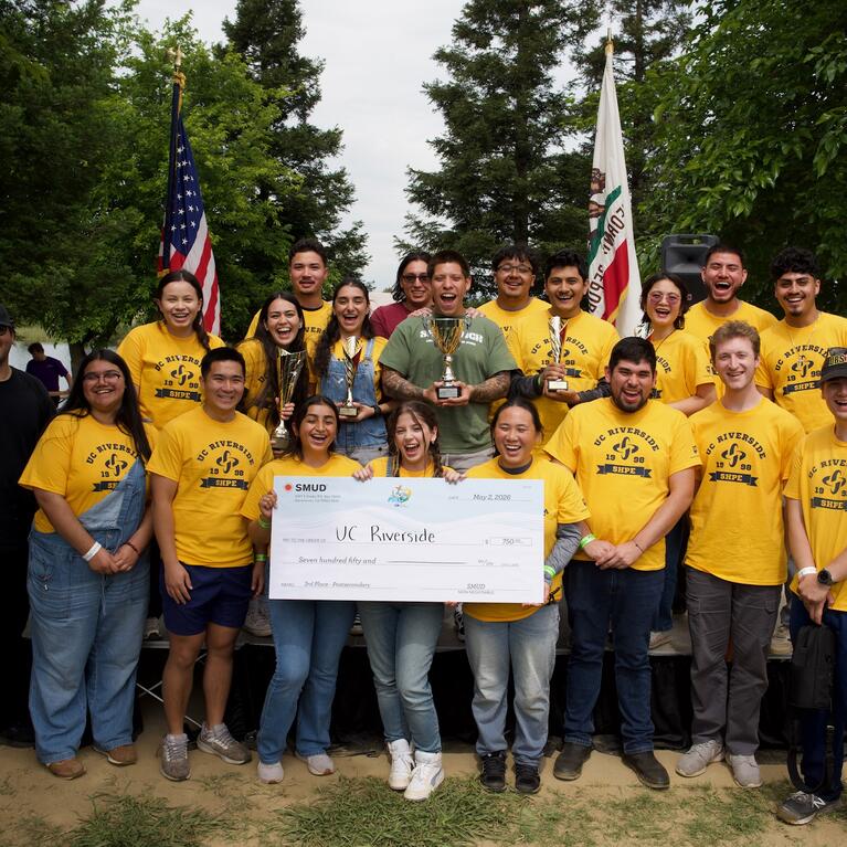 A group of UC Riverside students wearing matching yellow SHPE shirts stand outdoors together smiling and celebrating while holding trophies and an large award check made out to UCR. American and California state flags stand behind the group, with trees and a lakeside campsite visible in the background.