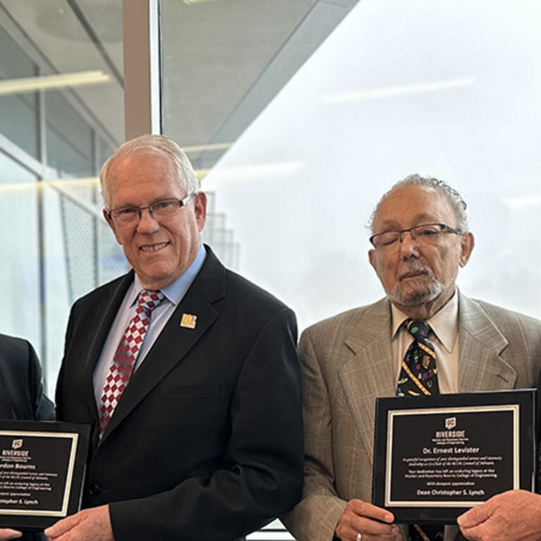 Four men wearing suits and ties are standing side by side, looking at the camera and smiling. From left to right:  Reza Abbaschian and Gordon Bourns hold a plaque, followed by Ernest Levister and Chris Lynch, also holding a plaque. The background features large windows with an outdoor view of trees and what appears to be a building in the distance.