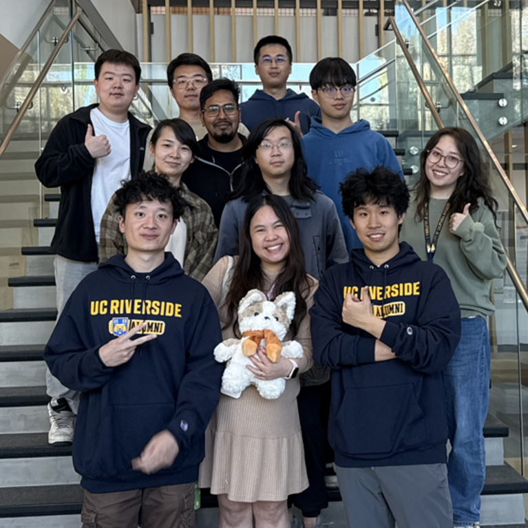Group of ten UC Riverside students posing on a staircase inside a campus building. Several students wear UCR hoodies, while one student in the center holds a plush mascot. The group smiles toward the camera with glass railings and a modern atrium visible in the background.