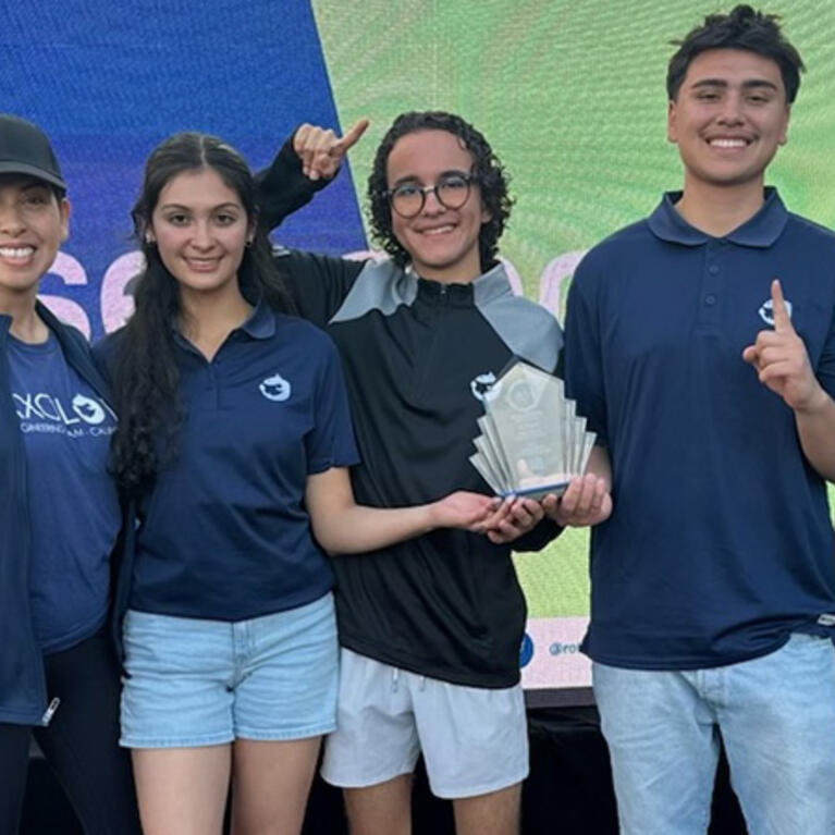 A diverse group of four students and MESA director standing together on a platform outdoors, holding a trophy and gesturing excitedly.