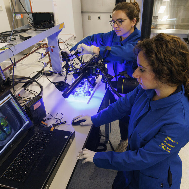 Two researchers in blue lab coats operating imaging and laboratory equipment at a computer workstation.