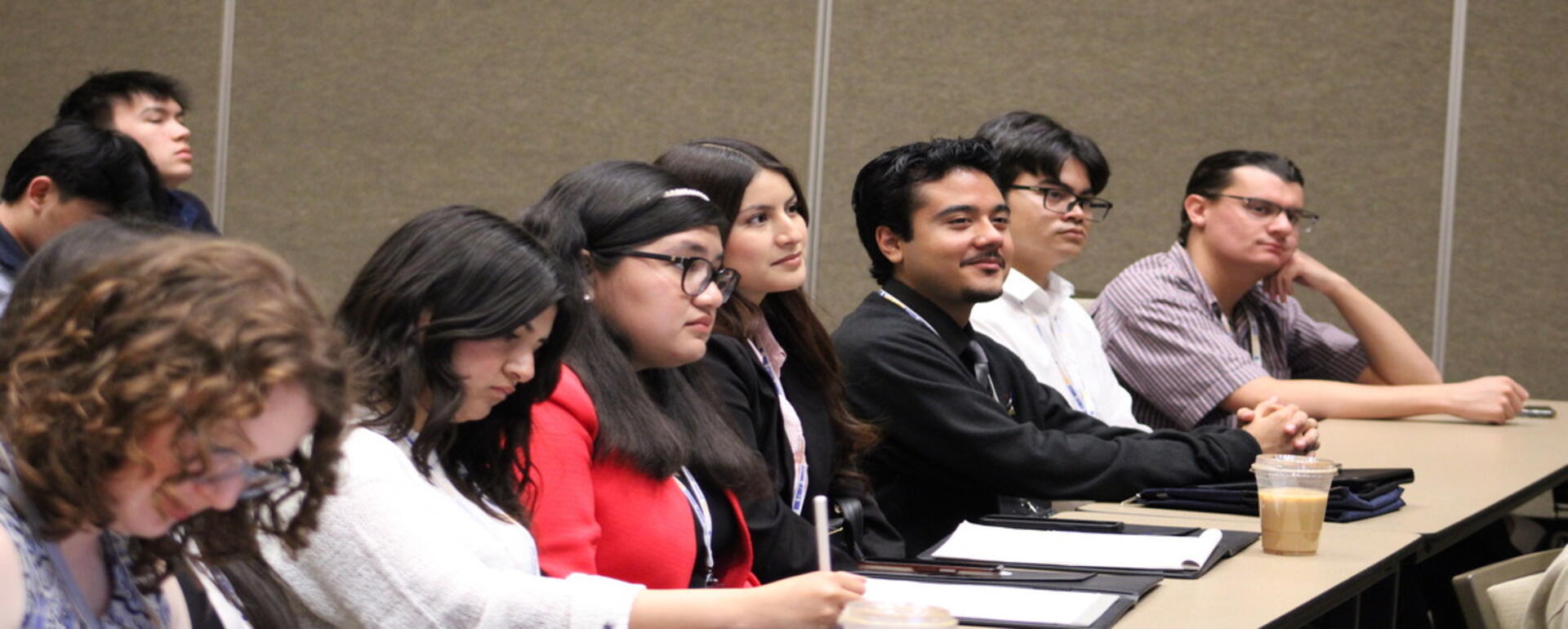 Several students wearing conference badges sit at tables during a professional development or networking session. One student takes notes while others listen attentively, with folders and a coffee cup visible on the table in a conference room setting.