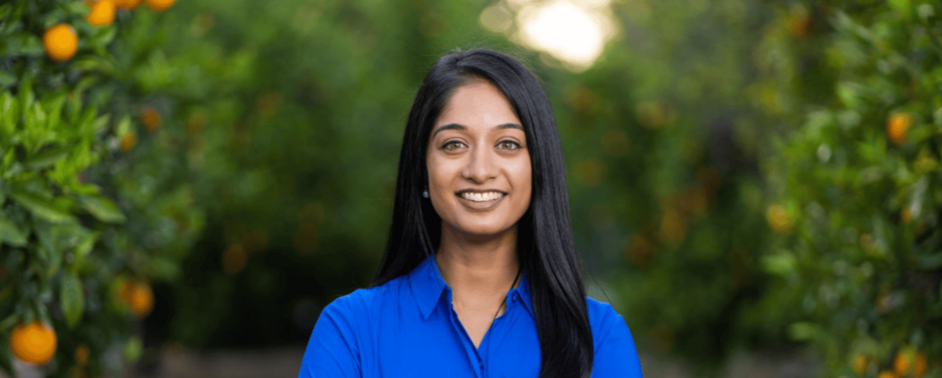 A woman wearing a black blazer and blue blouse stands outdoors for a professional portrait. She is smiling at the camera, with trees and a sunlit park-like setting softly blurred in the background.