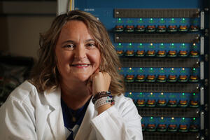 A woman wearing a white laboratory coat smiles at the camera while seated in front of rows of illuminated scientific equipment and electronic testing devices in a research laboratory.