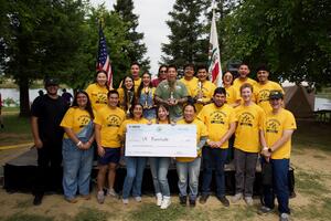 A group of UC Riverside students wearing matching yellow SHPE shirts stand outdoors together smiling and celebrating while holding trophies and an large award check made out to UCR. American and California state flags stand behind the group, with trees and a lakeside campsite visible in the background.
