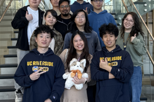 Group of ten UC Riverside students posing on a staircase inside a campus building. Several students wear UCR hoodies, while one student in the center holds a plush mascot. The group smiles toward the camera with glass railings and a modern atrium visible in the background.