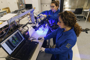 Two researchers in blue lab coats operating imaging and laboratory equipment at a computer workstation.