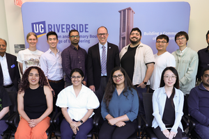 A group photo of thirteen individuals at UC Riverside, posed in front of a blue backdrop featuring the university logo and the Bell Tower. The group is arranged in two rows: six people are seated in the front row, and seven people are standing in the back row. They are dressed in a mix of professional and business-casual attire, including button-down shirts, sweaters, and blouses. The setting appears to be a formal academic seminar or engineering event, with a sign on the backdrop that reads "Building a Com