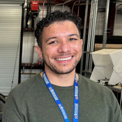 Headshot of Justin Bautista, a man with dark curly hair and a beard, smiling. He wears a green long-sleeved shirt and a blue lanyard.