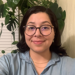 Vanessa Coria, a woman with dark hair and glasses, smiling faintly at the camera. She is wearing a light blue collared shirt and two delicate gold necklaces. Behind her, a large green plant is visible on the left, and white blinds are visible on the far left.