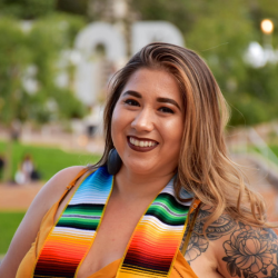 Yvette Casillas has long, light brown hair with highlights, dark eyes, and is wearing dark lipstick. She is wearing an orange top and a colorful striped stole.