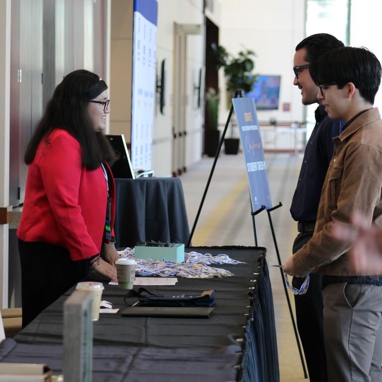 Woman with long brown hair, wearing reading glasses and a red blazer, speaking with two brown-haired men wearing glasses. 