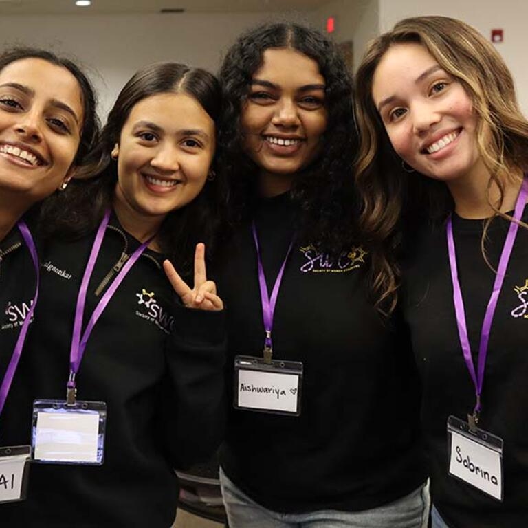 four women members wearing black t-shirts and purple lanyards smiling at the photographer 
