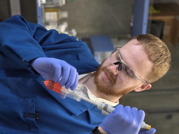 Jacob pipettes a red liquid into a centrifuge tube while wearing safety glasses, blue gloves, and a lab coat in a research laboratory.
