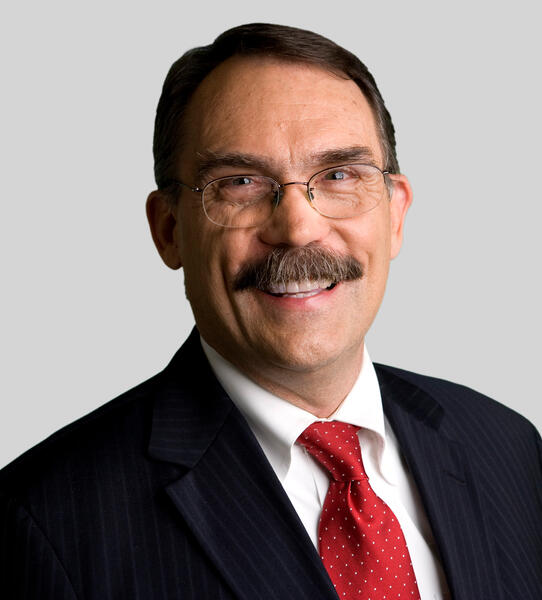 Professional headshot of a smiling man wearing glasses, a dark pinstripe suit, white dress shirt, and red patterned tie against a light gray background. He is facing the camera and appears in a formal business portrait.