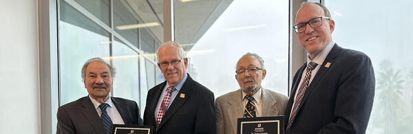 Four men wearing suits and ties are standing side by side, looking at the camera and smiling. From left to right:  Reza Abbaschian and Gordon Bourns hold a plaque, followed by Ernest Levister and Chris Lynch, also holding a plaque. The background features large windows with an outdoor view of trees and what appears to be a building in the distance.