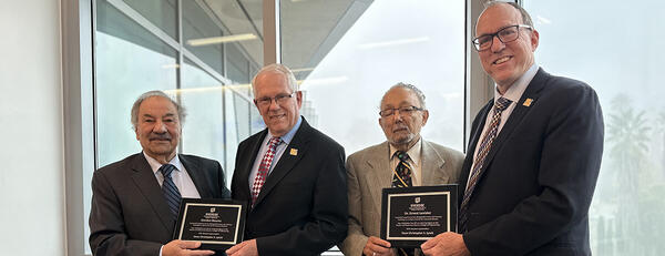 Four men wearing suits and ties are standing side by side, looking at the camera and smiling. From left to right:  Reza Abbaschian and Gordon Bourns hold a plaque, followed by Ernest Levister and Chris Lynch, also holding a plaque. The background features large windows with an outdoor view of trees and what appears to be a building in the distance.