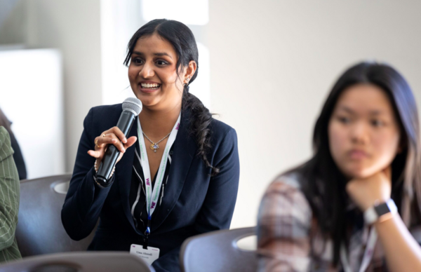 Woman in the foreground holding a microphone wearing a black blazer and white blouse. Women in the background in a plaid shirt resting her chin on her knuckles.  