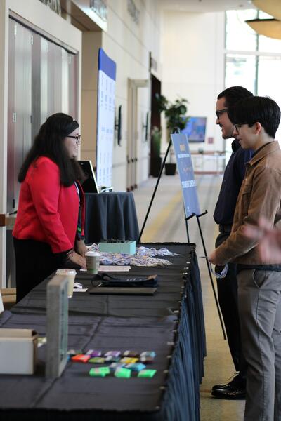 Woman with long brown hair, wearing reading glasses and a red blazer, speaking with two brown-haired men wearing glasses. 