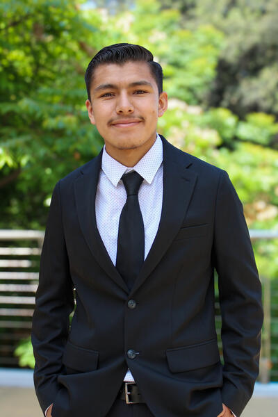 Joe Jimenez, an early twenties Hispanic male wearing black suit and tie with a white shirt smiling