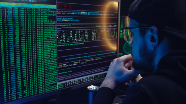 bearded man with cap and glasses looking at multiple monitors with dark screens and colorful numbers and characters 