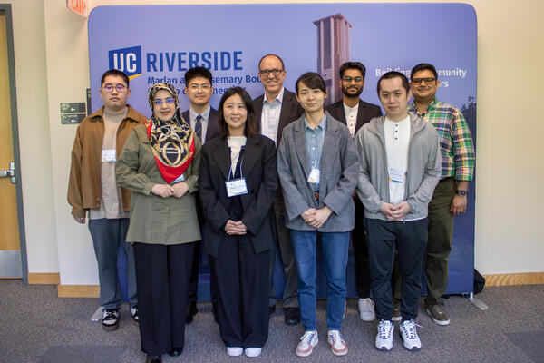 Nine people in business casual attire posing indoors in front of a UC Riverside backdrop, smiling at the camera