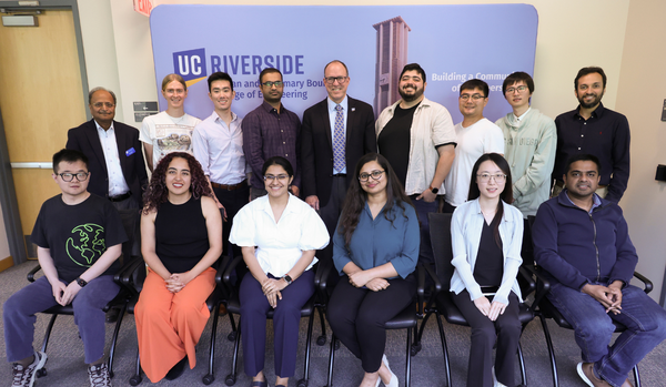 A group photo of thirteen individuals at UC Riverside, posed in front of a blue backdrop featuring the university logo and the Bell Tower. The group is arranged in two rows: six people are seated in the front row, and seven people are standing in the back row. They are dressed in a mix of professional and business-casual attire, including button-down shirts, sweaters, and blouses. The setting appears to be a formal academic seminar or engineering event, with a sign on the backdrop that reads "Building a Com