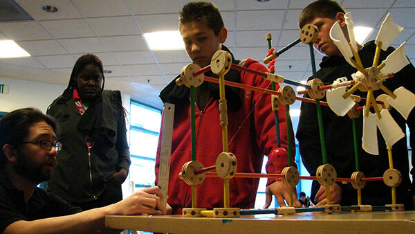 A MESA instructor guides three students who are gathered around a table through a turbine-model-making session during an engineering competition.