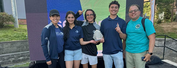 A diverse group of four students and MESA director standing together on a platform outdoors, holding a trophy and gesturing excitedly.