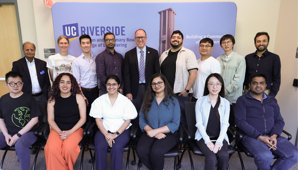A group photo of thirteen individuals at UC Riverside, posed in front of a blue backdrop featuring the university logo and the Bell Tower. The group is arranged in two rows: six people are seated in the front row, and seven people are standing in the back row. They are dressed in a mix of professional and business-casual attire, including button-down shirts, sweaters, and blouses. The setting appears to be a formal academic seminar or engineering event, with a sign on the backdrop that reads "Building a Com