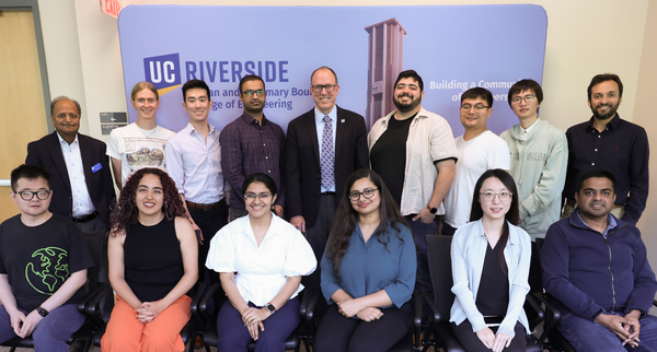 A group photo of thirteen individuals at UC Riverside, posed in front of a blue backdrop featuring the university logo and the Bell Tower. The group is arranged in two rows: six people are seated in the front row, and seven people are standing in the back row. They are dressed in a mix of professional and business-casual attire, including button-down shirts, sweaters, and blouses. The setting appears to be a formal academic seminar or engineering event, with a sign on the backdrop that reads "Building a Com