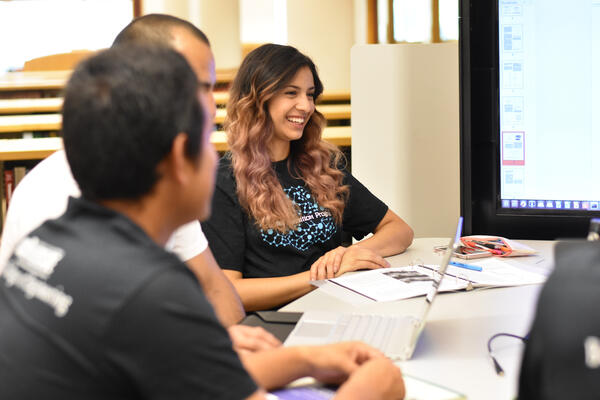 a young woman and two men sitting at a desk and looking ahead at monitors