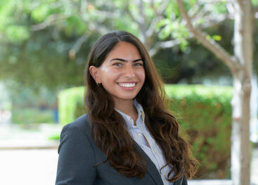 UC Riverside bioengineering student Janna Soliman stands outdoors smiling at the camera while wearing a dark blazer over a striped button-up shirt. Trees, greenery, and a sunlit walkway appear softly blurred in the background.