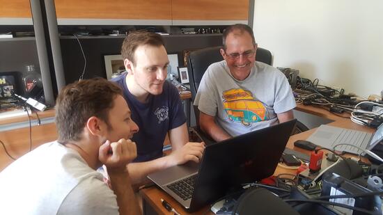 Three men gather around laptops and electronic equipment in an office while working together on a project.