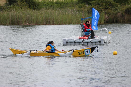 A UCR SHPE student wearing a life jacket pilots a yellow solar-powered boat across a lake during the SMUD Solar Regatta, while race officials observe from a floating dock in the background.
