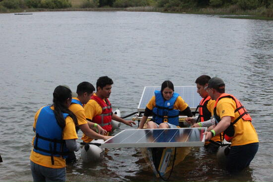 Members of the UCR SHPE Solar Boat team stand in shallow water wearing life jackets as they guide a solar-powered boat equipped with large solar panels during a regatta competition at a lakeside course.