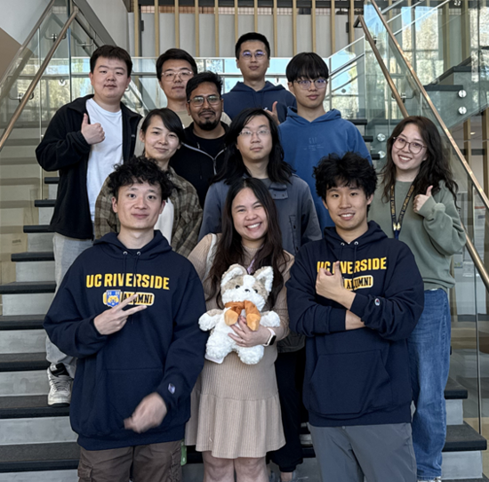 Group of ten UC Riverside students posing on a staircase inside a campus building. Several students wear UCR hoodies, while one student in the center holds a plush mascot. The group smiles toward the camera with glass railings and a modern atrium visible in the background.