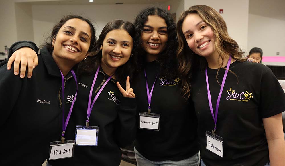 four women members wearing black t-shirts and purple lanyards smiling at the photographer 